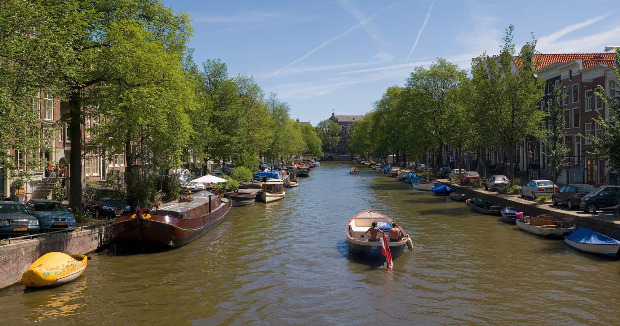 Un canal din Amsterdam vara, cu bărci și copaci pe cheiuri.