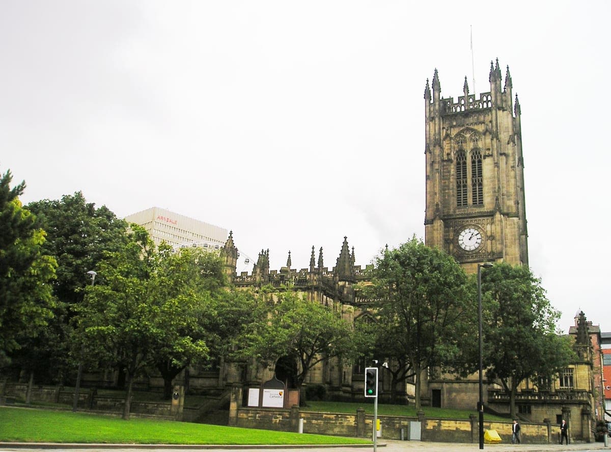 Catedrala din Manchester văzută de pe stradă, cu turnul cu ceas deasupra copacilor.